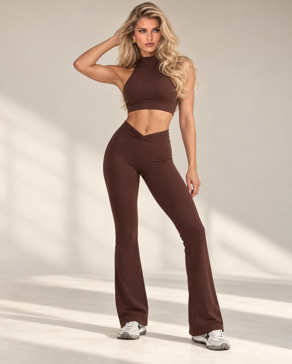 ERAFITT women's chocolate brown matching activewear set featuring a sleeveless high-neck cropped athletic top with a fitted body-contouring silhouette, wide underbust band and coordinating high-waisted full-length flared leggings. The rich brown two-piece workout outfit features smooth stretch-look fabric, a flattering V-shaped crossover waistband, sculpted waist and hip fit, fitted thighs and softly flared hems. Blonde model showcasing the front design of the matching brown gym outfit with white athletic s