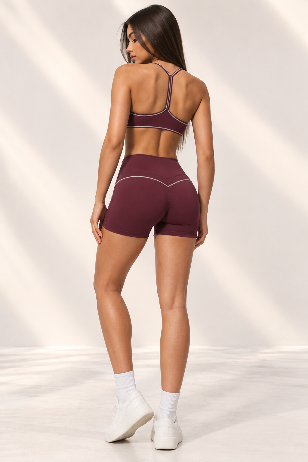 burgundy women's workout set combining a sleek cropped sports bra with curved lower edge and high-waisted fitted shorts, photographed in a warm cream studio.